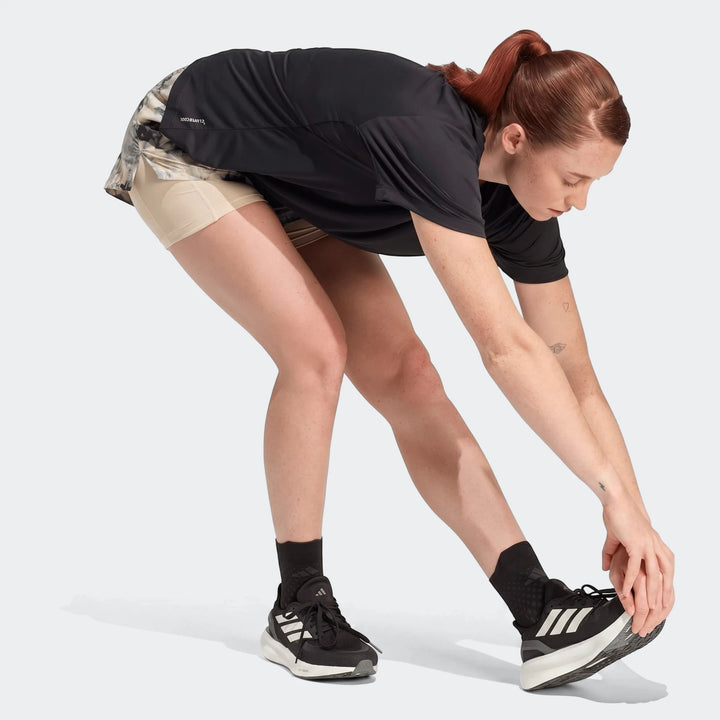Woman in a black Run Essentials T-shirt and patterned running shorts stretching. She is wearing black and white athletic