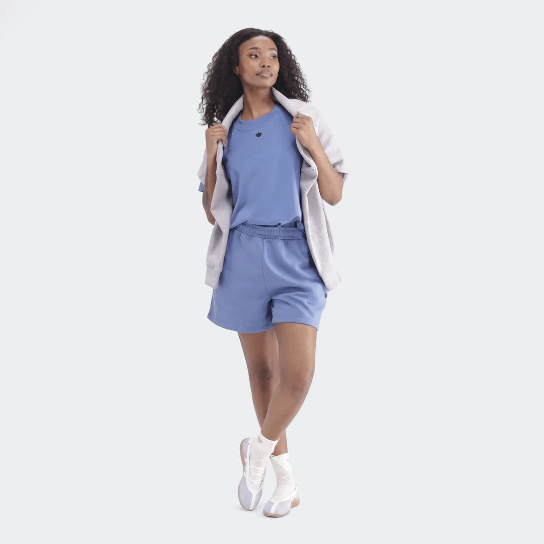 Full-body shot of a woman wearing a blue Essentials Trefoil T-Shirt, blue shorts, and white sneakers, looking right.