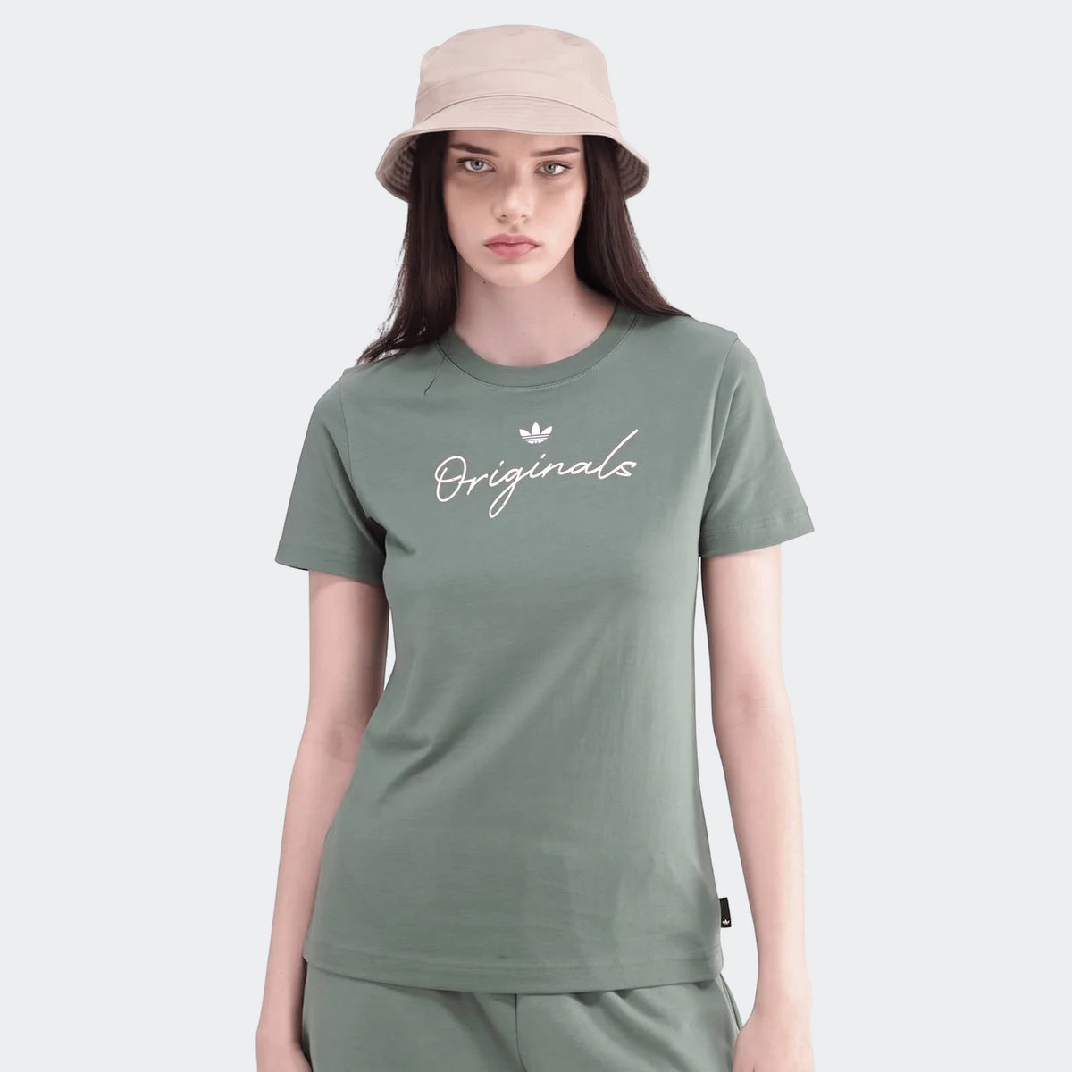 Woman wearing a green Adidas Originals short sleeve t-shirt, matching green pants, and a beige bucket hat, looking directly