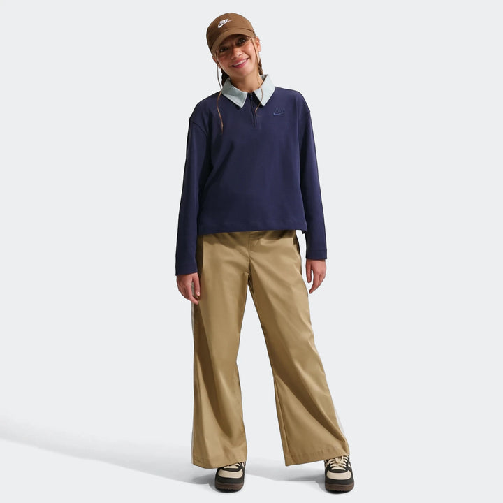 Woman wearing a brown Nike cap, a navy Nike long-sleeve polo shirt, and parachute brown pants, posing in a full body shot.