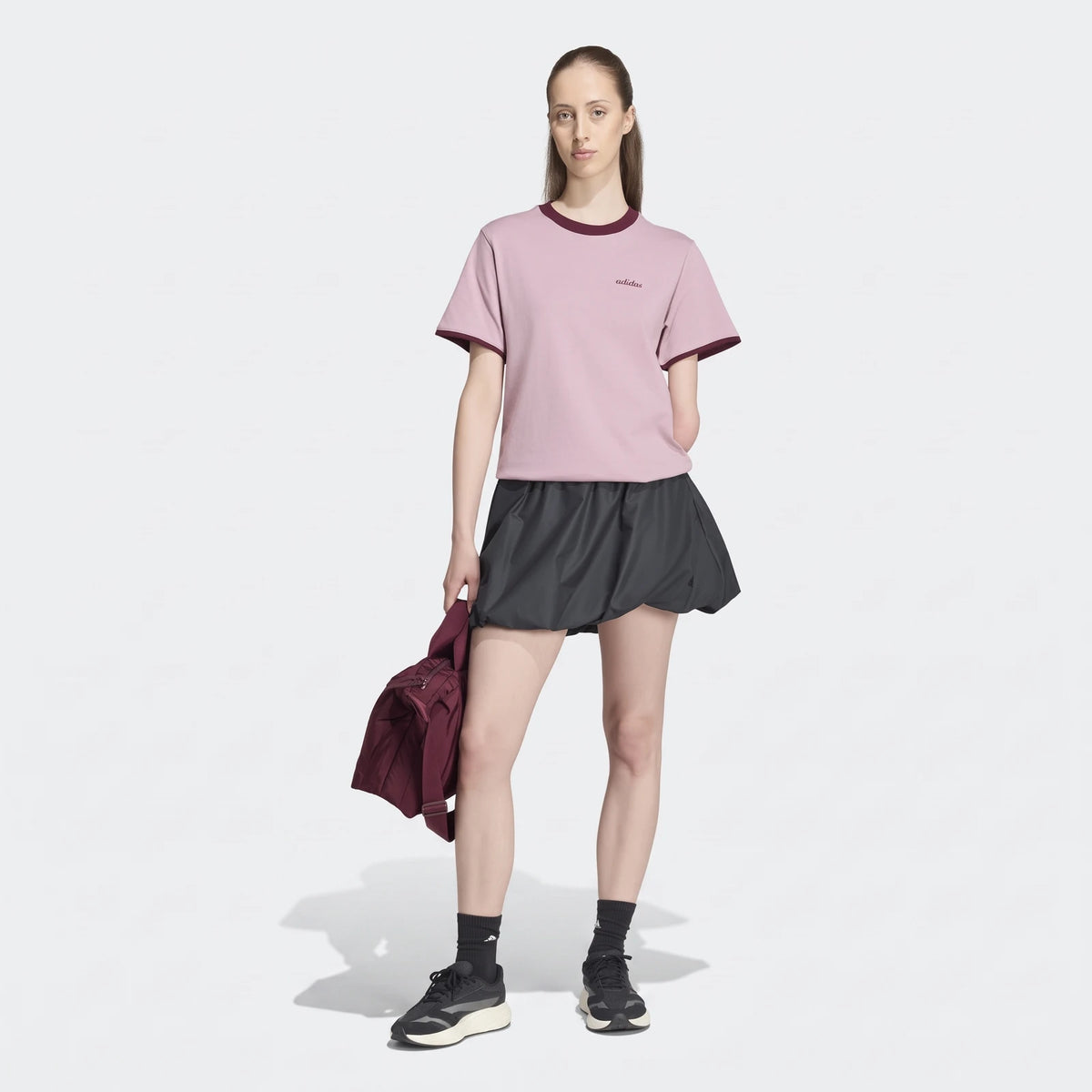Woman wearing a pink adidas Seasonal Essentials Colorpop T-Shirt with an embroidered graphic, dark skirt, and maroon bag.