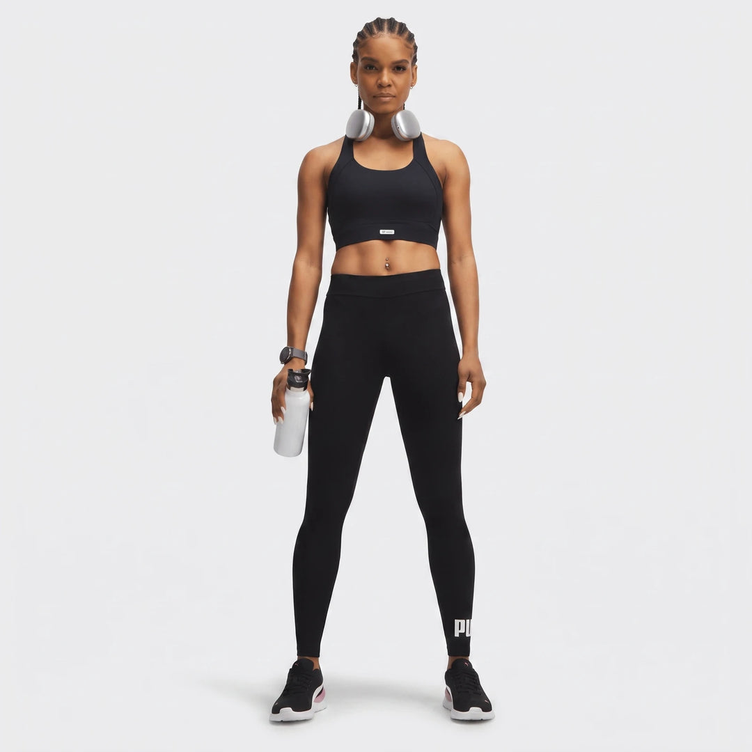 Woman in black PUMA Ess No:1 Logo Leggings, black sports bra, and silver headphones, holding a white water bottle.