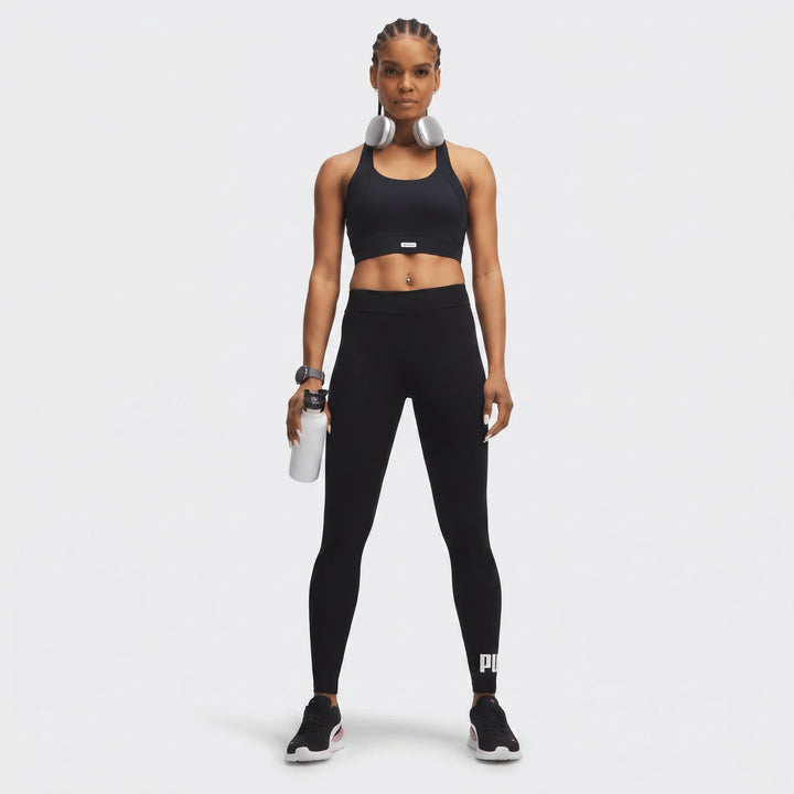 Woman in black PUMA Ess No:1 Logo Leggings, black sports bra, and silver headphones, holding a white water bottle.