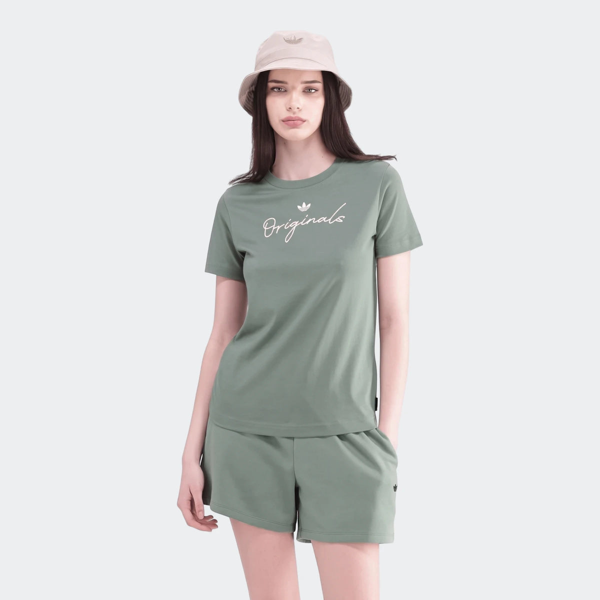 Woman models a sage green short-sleeve t-shirt with an 'Originals' logo, paired with matching shorts and a beige bucket hat.