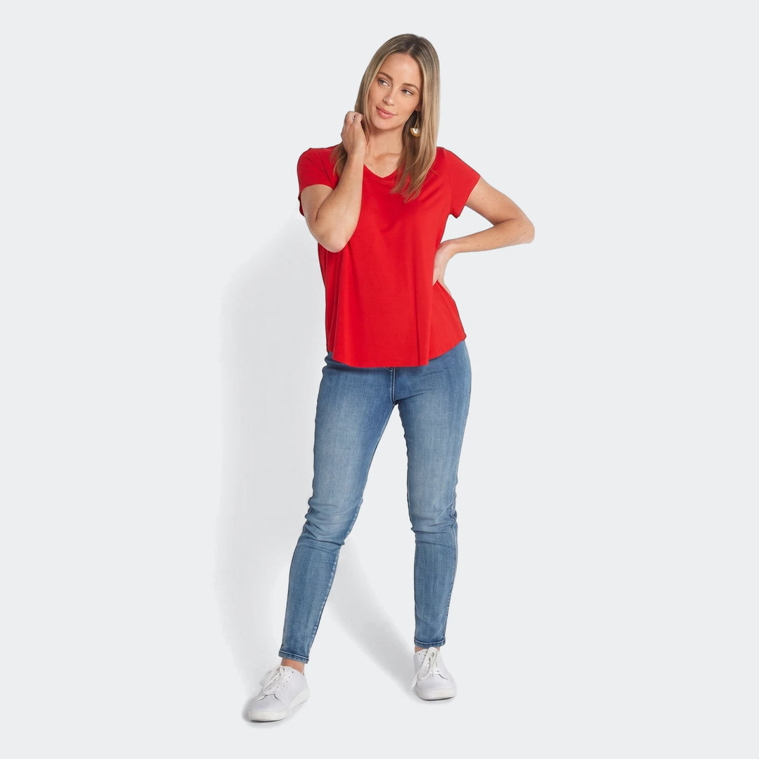 Woman wearing a red V-neck t-shirt with blue jeans and white sneakers, standing full-body in a studio setting.