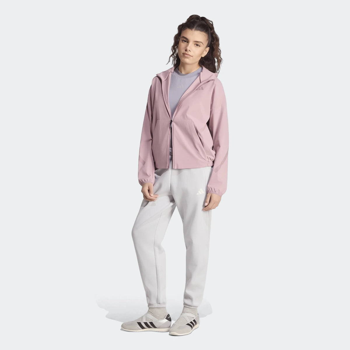 A female model wearing a pink Adidas Z.N.E. Woven Track Top, light gray sweatpants, and sneakers in a full body shot.