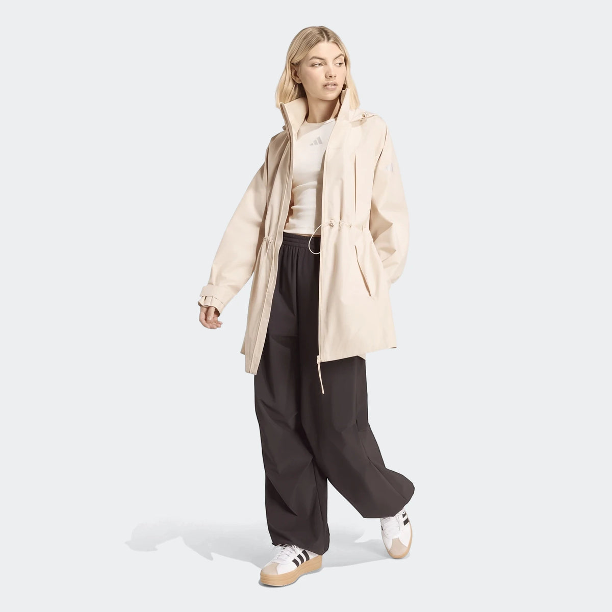 A blonde woman models a beige women's City Rain Parka, black pants, and white sneakers in a full body shot.
