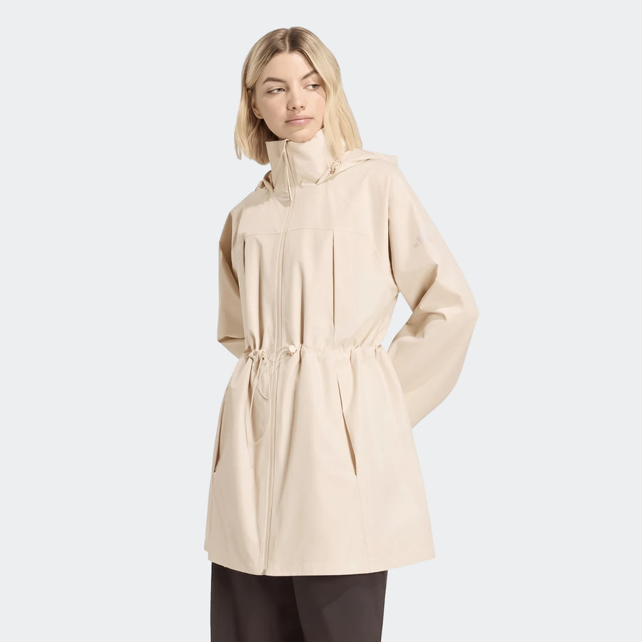 Blonde woman modeling a cream City Rain Parka with a hood, high collar, and drawstring waist on a white background, looking