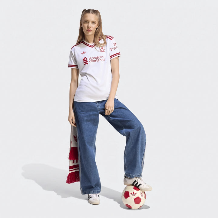 Woman wearing a white Liverpool FC 26/27 away jersey, blue jeans, and sneakers, with one foot on a soccer ball, holding a