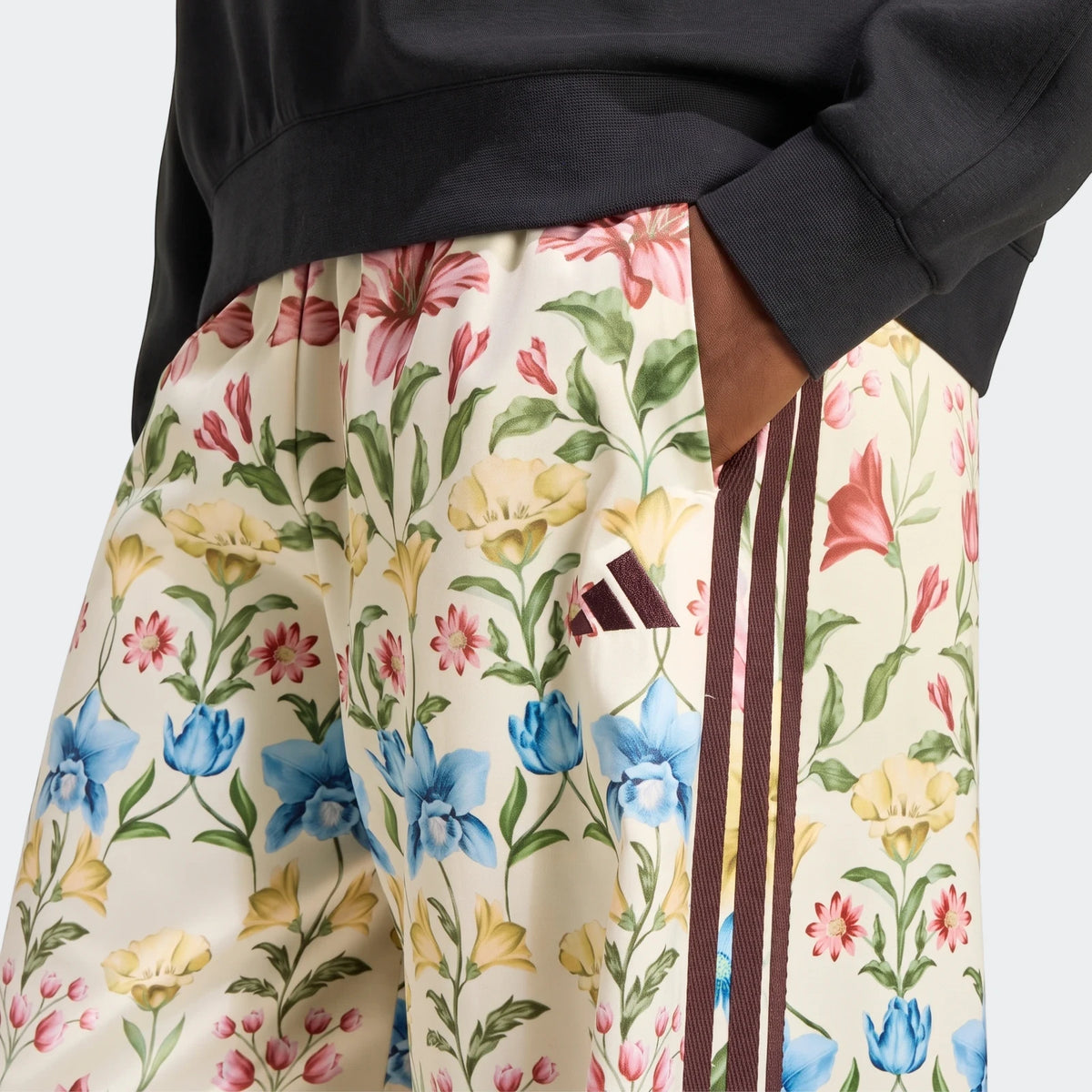 Close-up of X Farm Rio Premium Joggers in a cream floral pattern with brown Adidas stripes, worn by a person with their hand