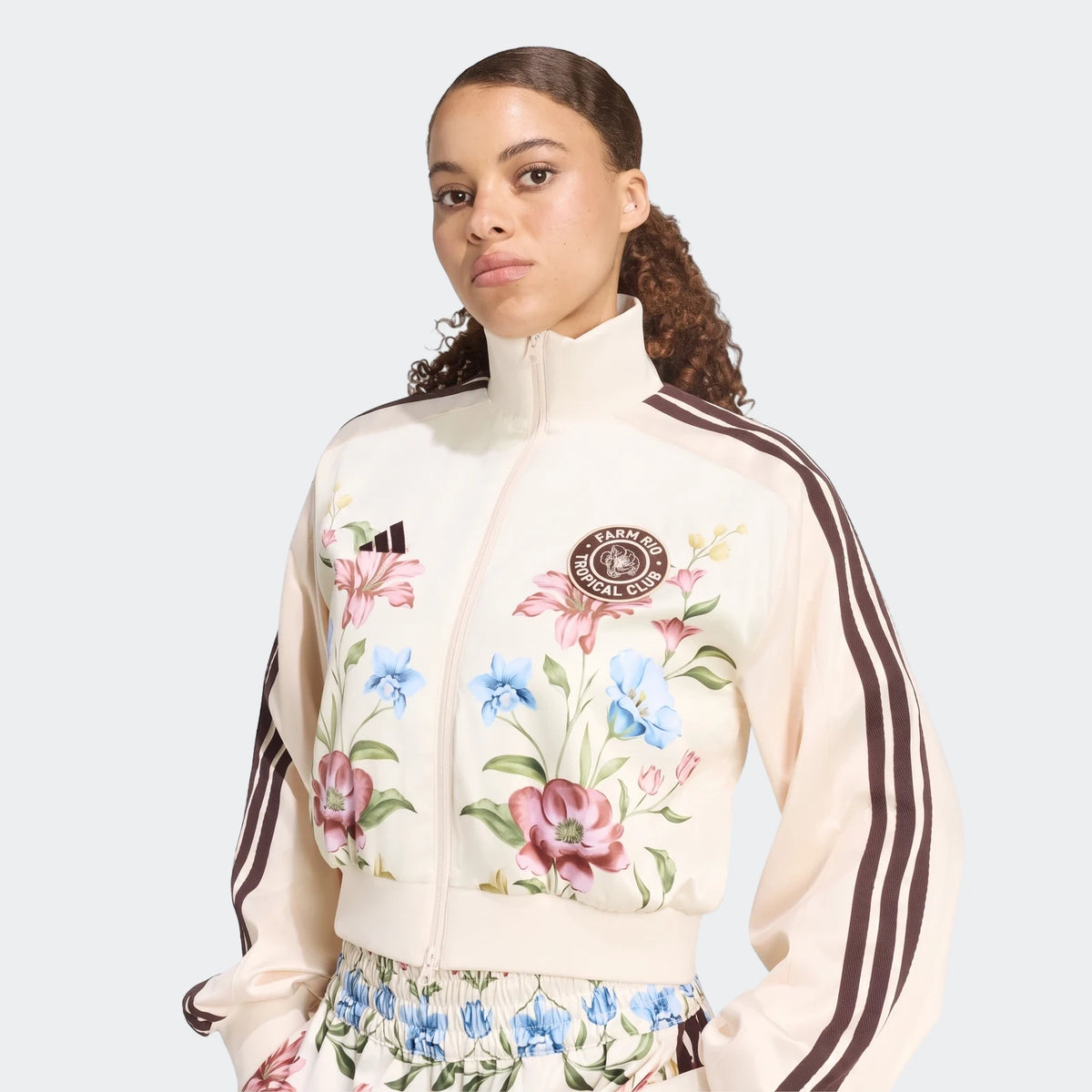 Waist-up view of a woman wearing a cream X Farm Rio Premium Tracktop featuring a vibrant floral print and iconic brown three