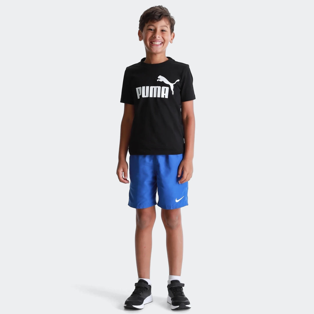 Young boy smiling, wearing a black Puma Ess Logo Tee T-shirt with white logo, blue shorts, and black sneakers.