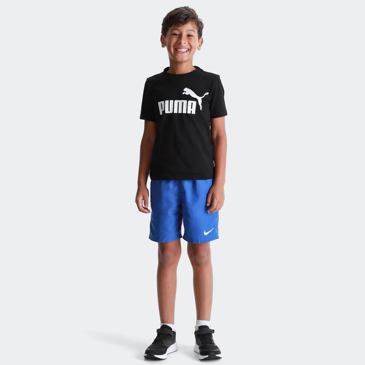 Young boy smiling, wearing a black Puma Ess Logo Tee T-shirt with white logo, blue shorts, and black sneakers.
