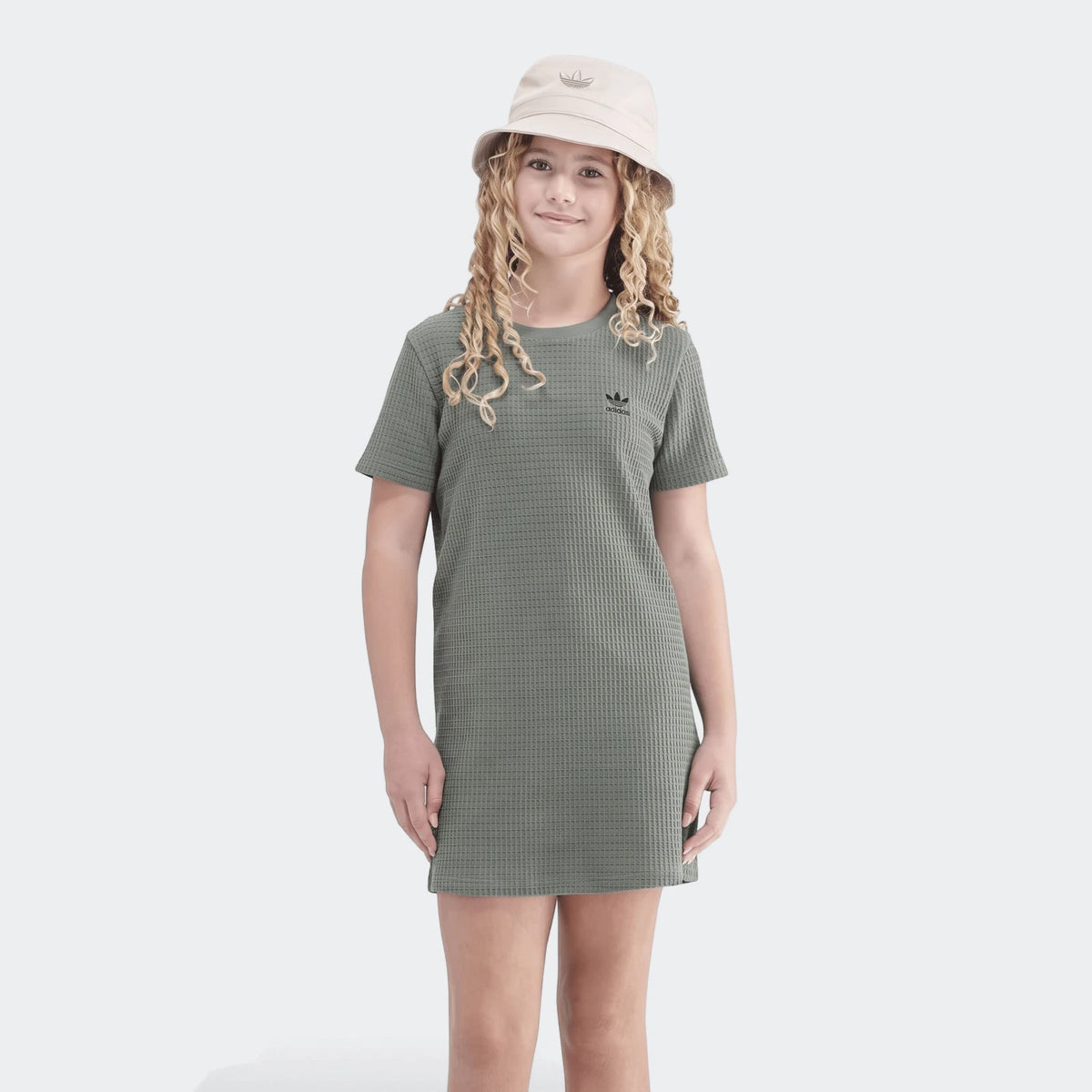 Young girl smiling in a green adidas waffle dress and beige bucket hat on a white background, full length.