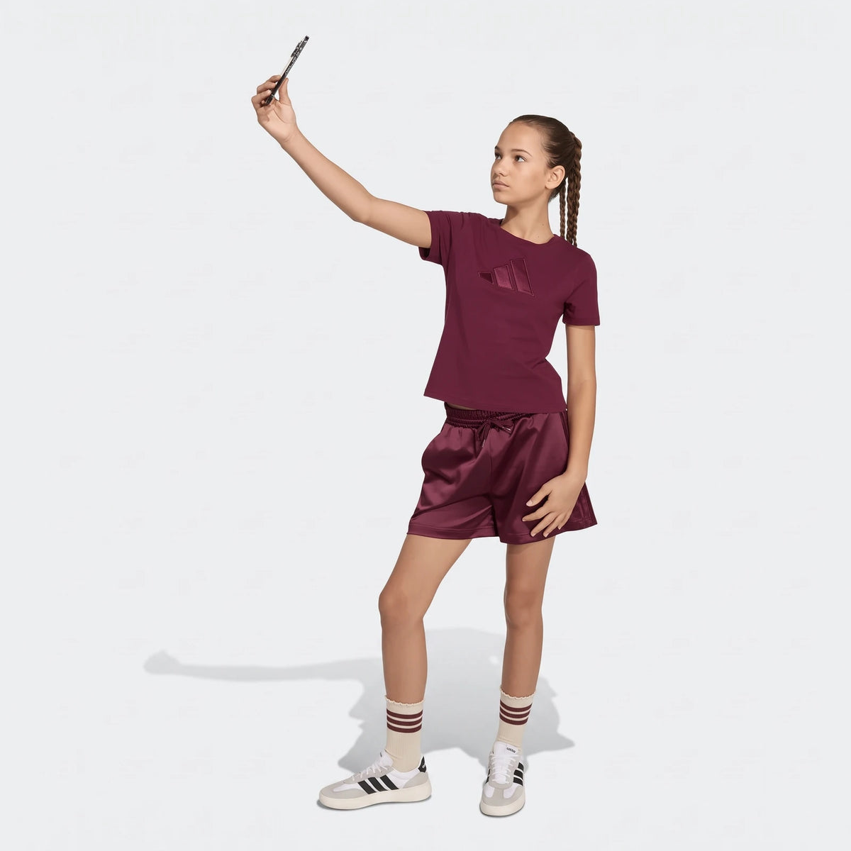 A young girl in a maroon Adidas t-shirt and matching satin shorts, with striped socks and white sneakers, taking a selfie.