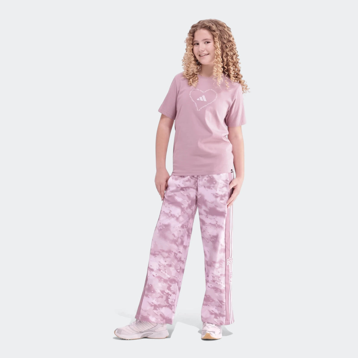 Smiling young girl with curly hair models a pink Adidas heart logo t-shirt and purple tie-dye wide-leg track pants with