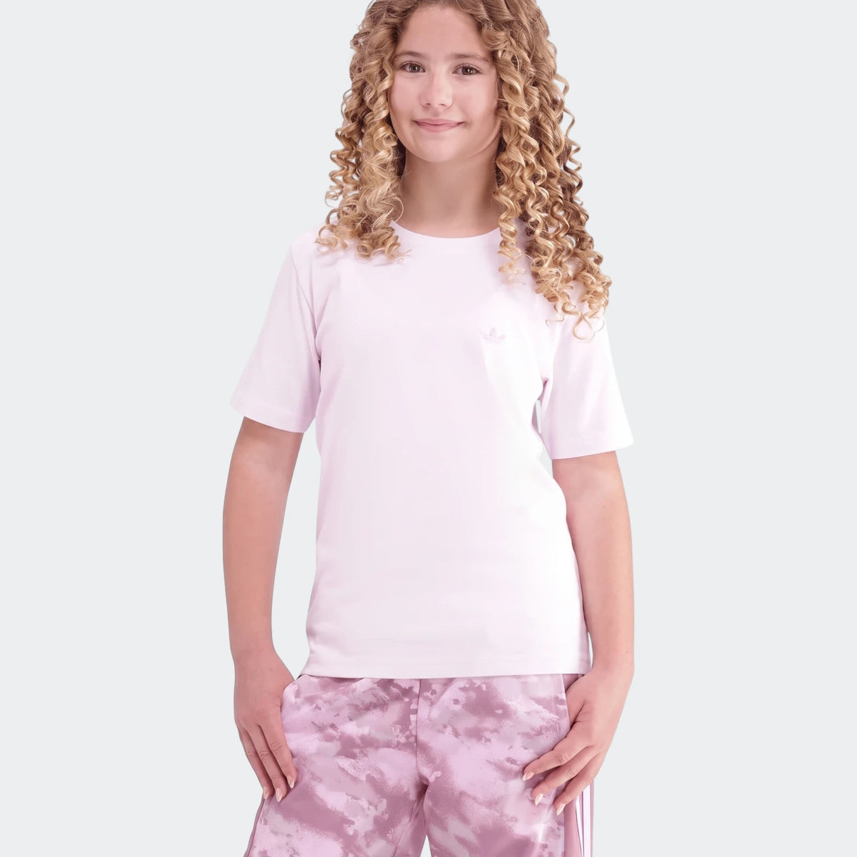Young girl with curly blonde hair wearing a light pink Adidas Premium Essentials Tee and pink patterned shorts, standing.