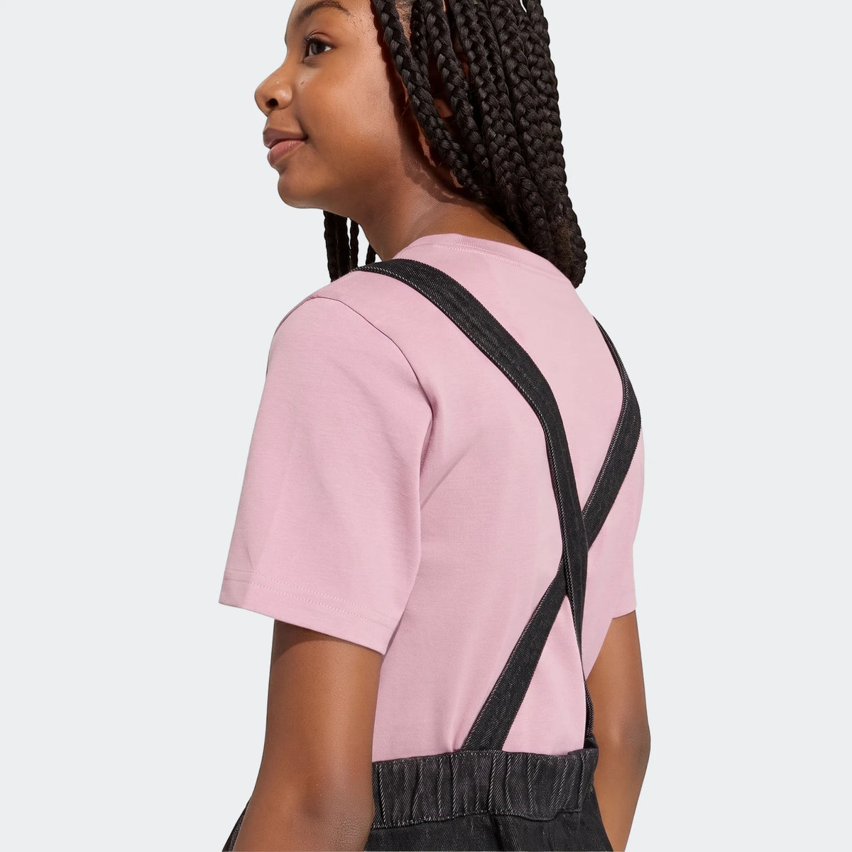 Young girl wearing a pink t-shirt and dark denim dungarees dress, seen from the back-left side with braided hair.