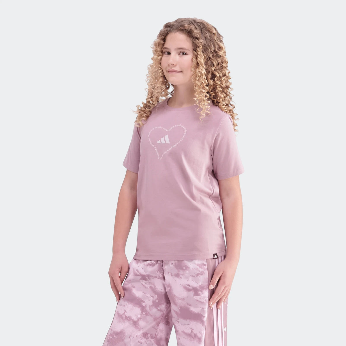 A young girl wearing a purple Adidas Jg Satin T-shirt with a white heart graphic and matching purple track pants with white