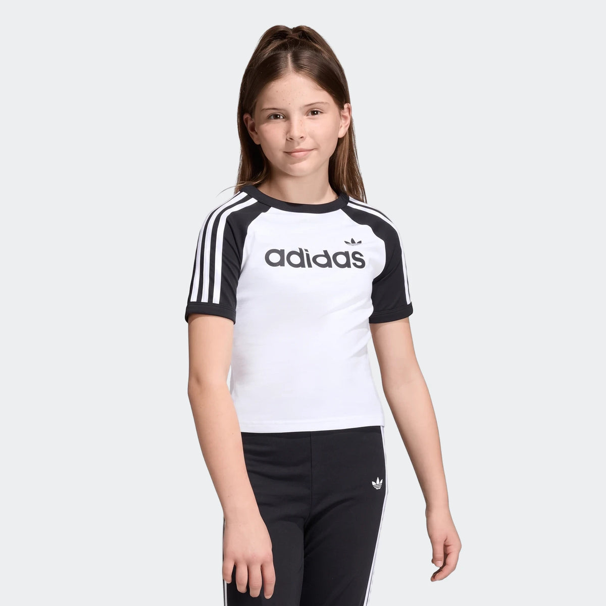 Young girl wearing a white Adidas 3 Stripes Slim T-Shirt with black sleeves and matching black track pants.