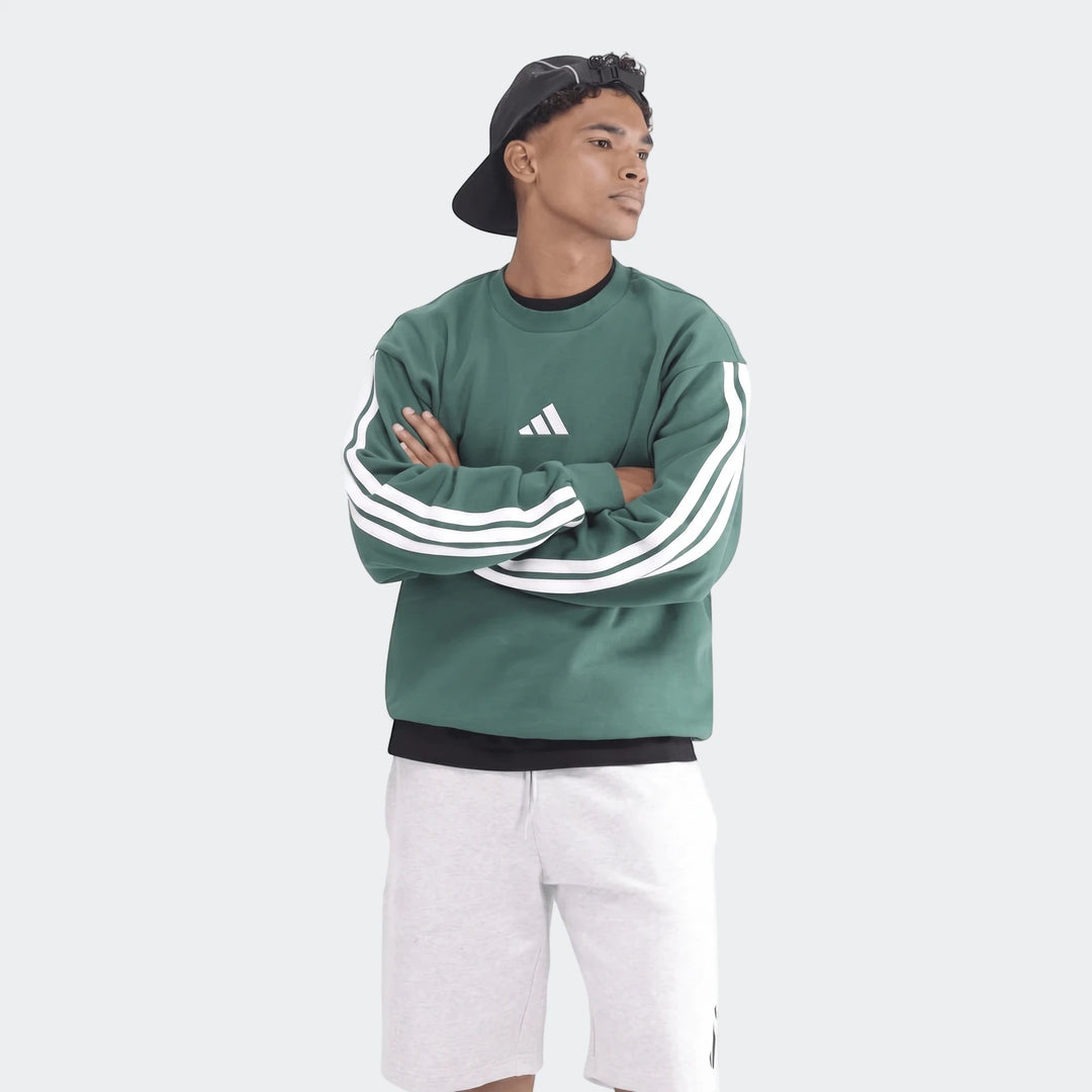 Young man wearing a green Adidas sweatshirt with white stripes, light gray shorts, and a black baseball cap. He stands with