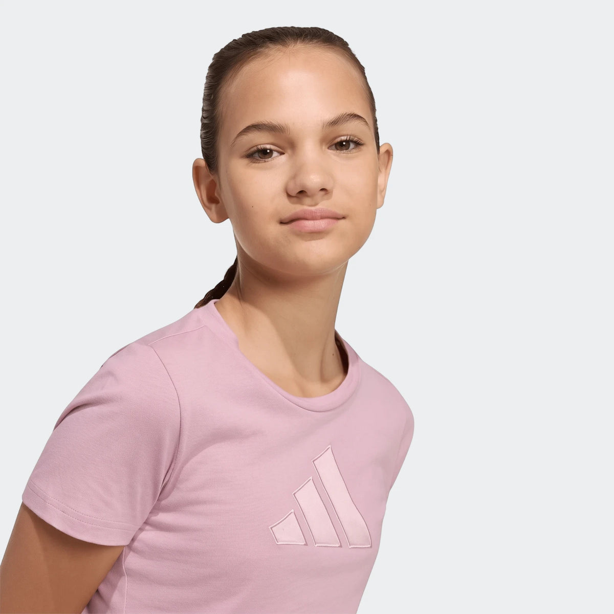 Young woman wearing a light pink Adidas Satin T-Shirt, cropped waist-up. The t-shirt features an embossed logo on the chest.