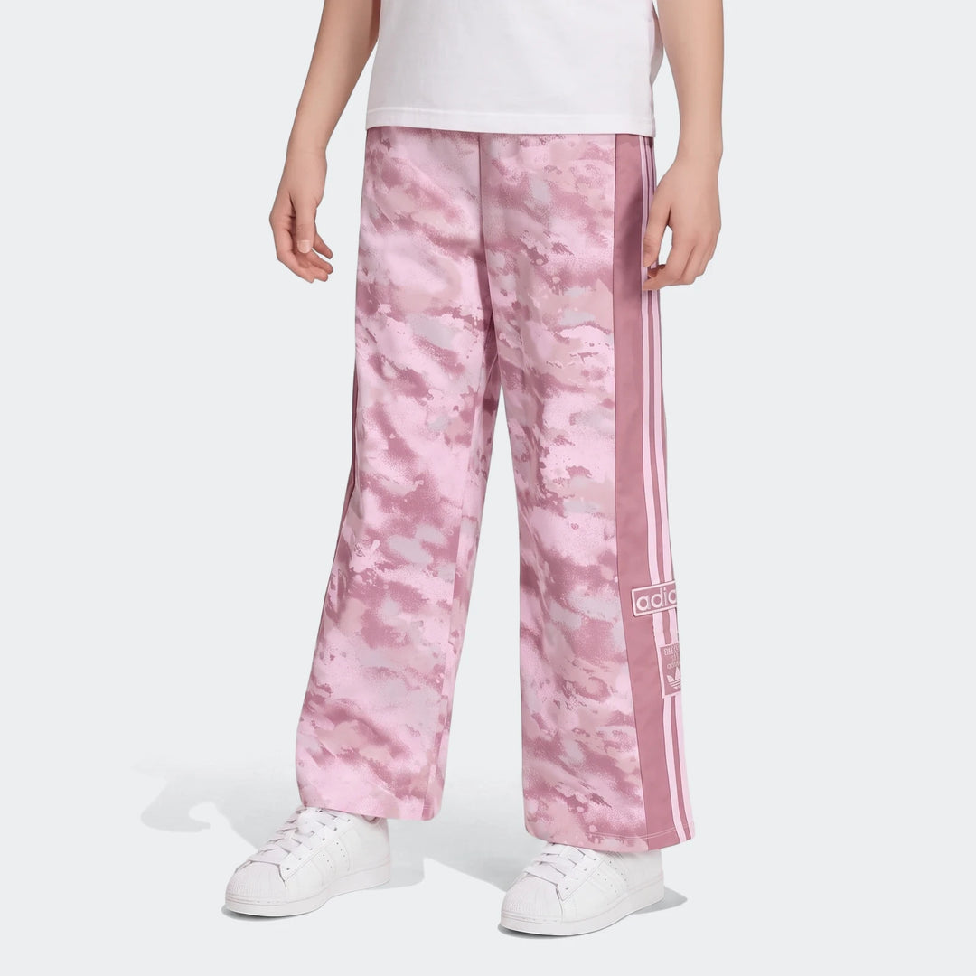 Pink camouflage Adidas Loose Adibreak twill trousers for youth, featuring white side stripes, worn with white sneakers.