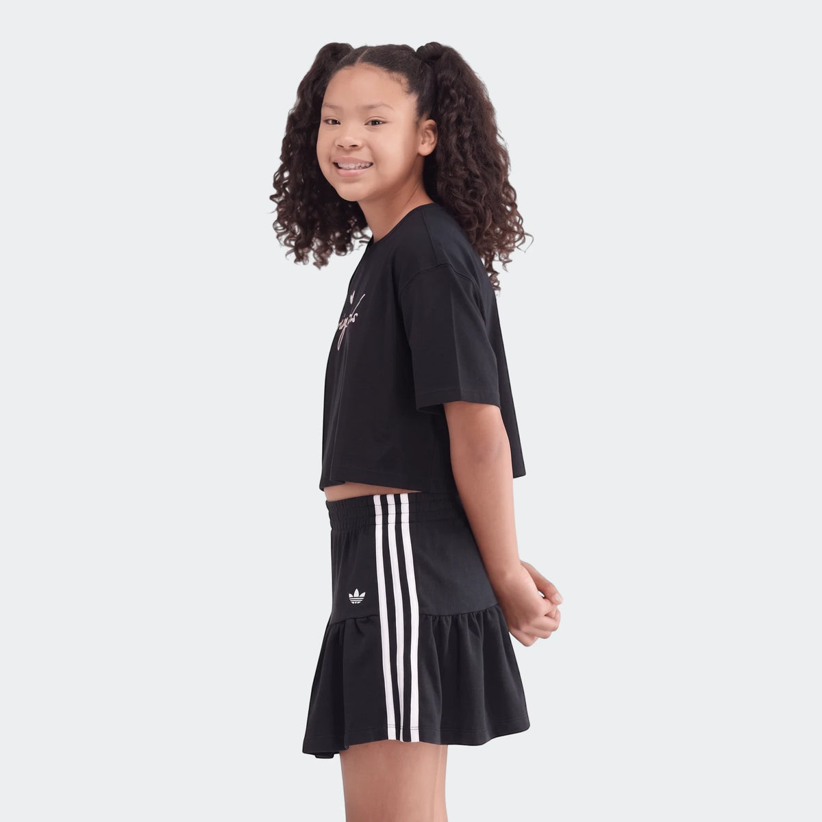 A young girl with curly hair smiles, wearing a black Adidas Youth Script Tee crop top and a matching black ruffled skirt