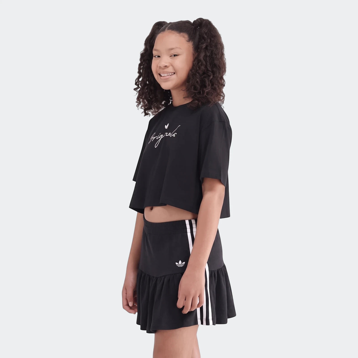 Young girl in a black Adidas Youth Script Tee and matching black ruffled skirt with white stripes, smiling and posing.