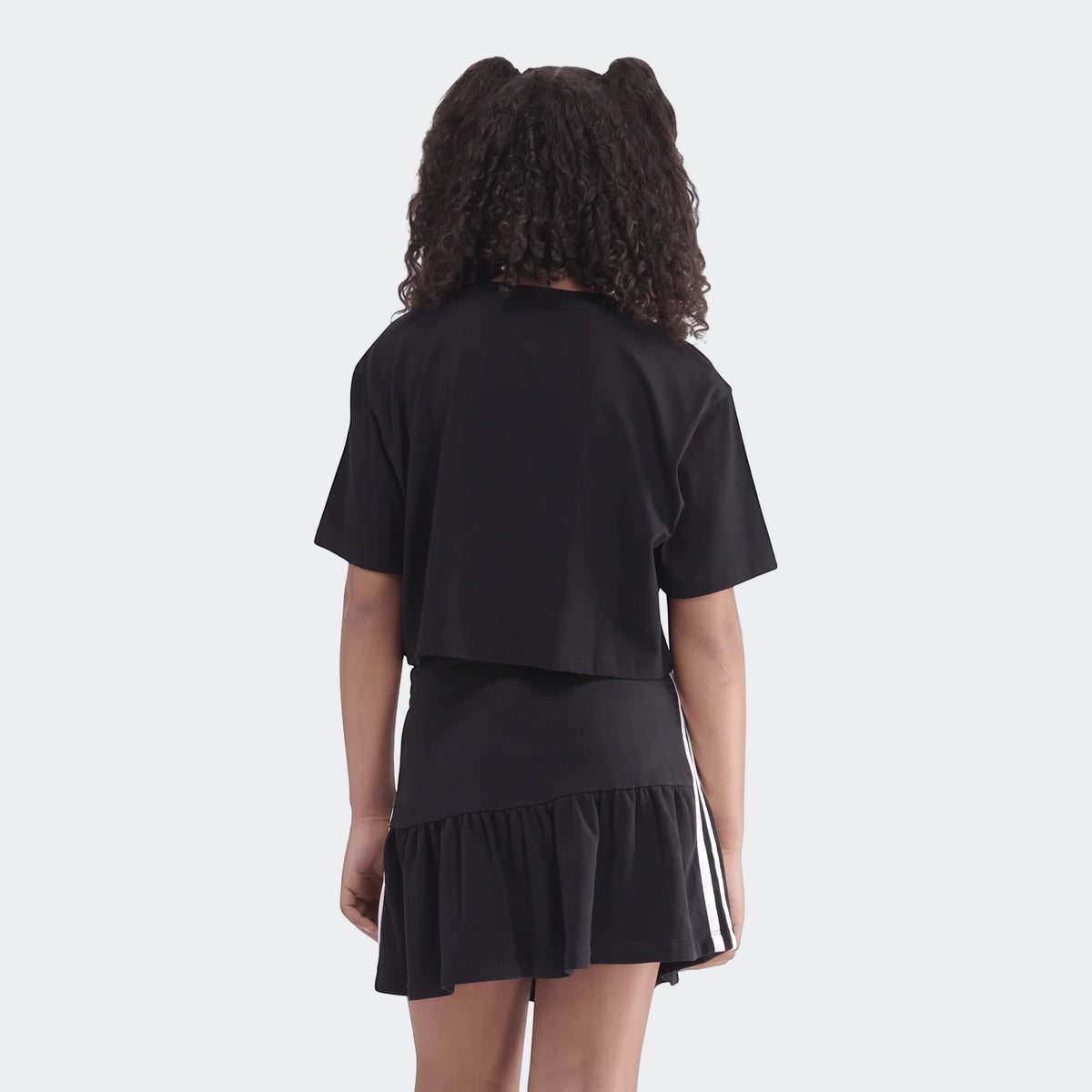 Youth Script Tee in black, worn by a girl facing away, paired with a black ruffled skirt featuring white side stripes.