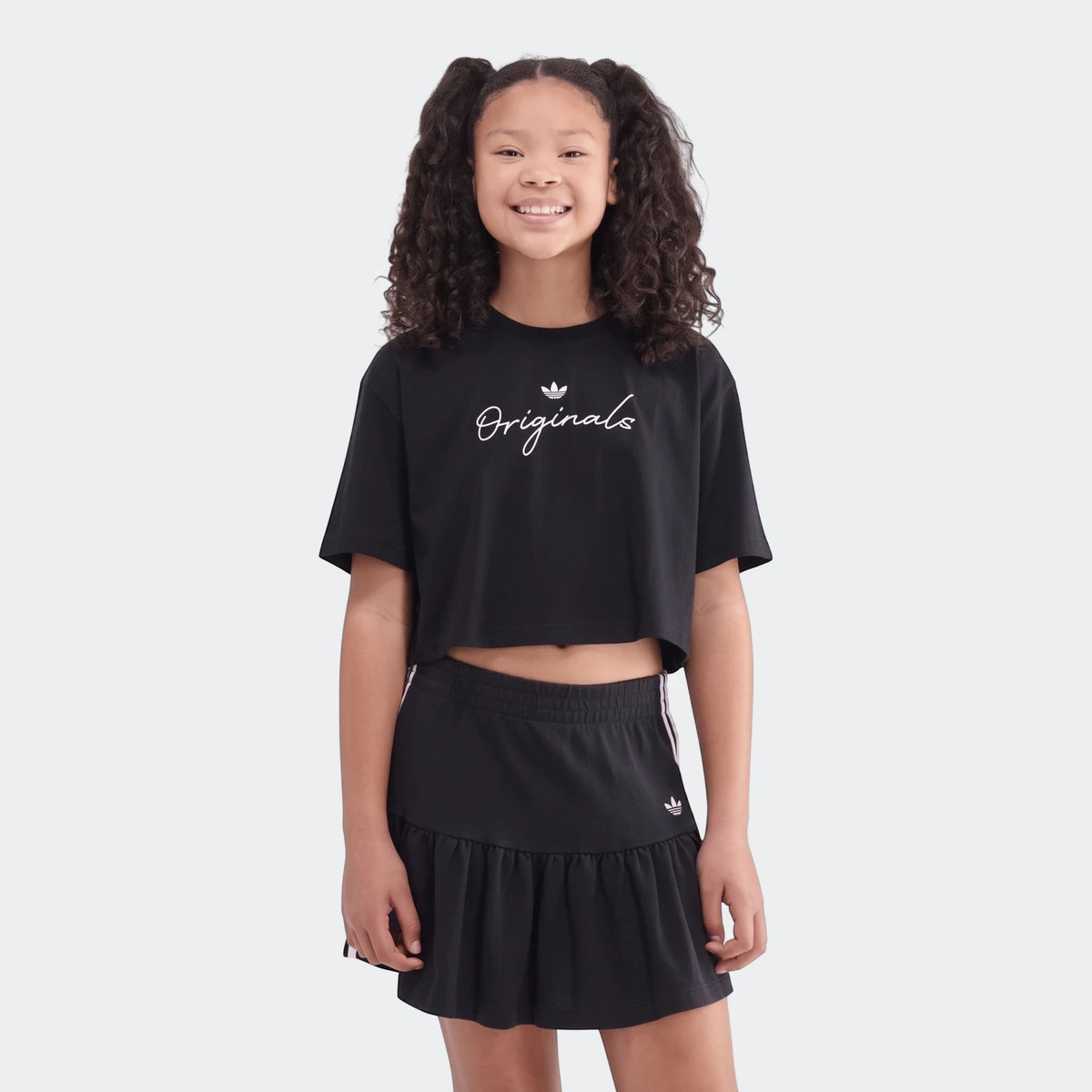 Young girl wearing a black Adidas Originals Youth Script Tee crop top and a black ruffled mini skirt.