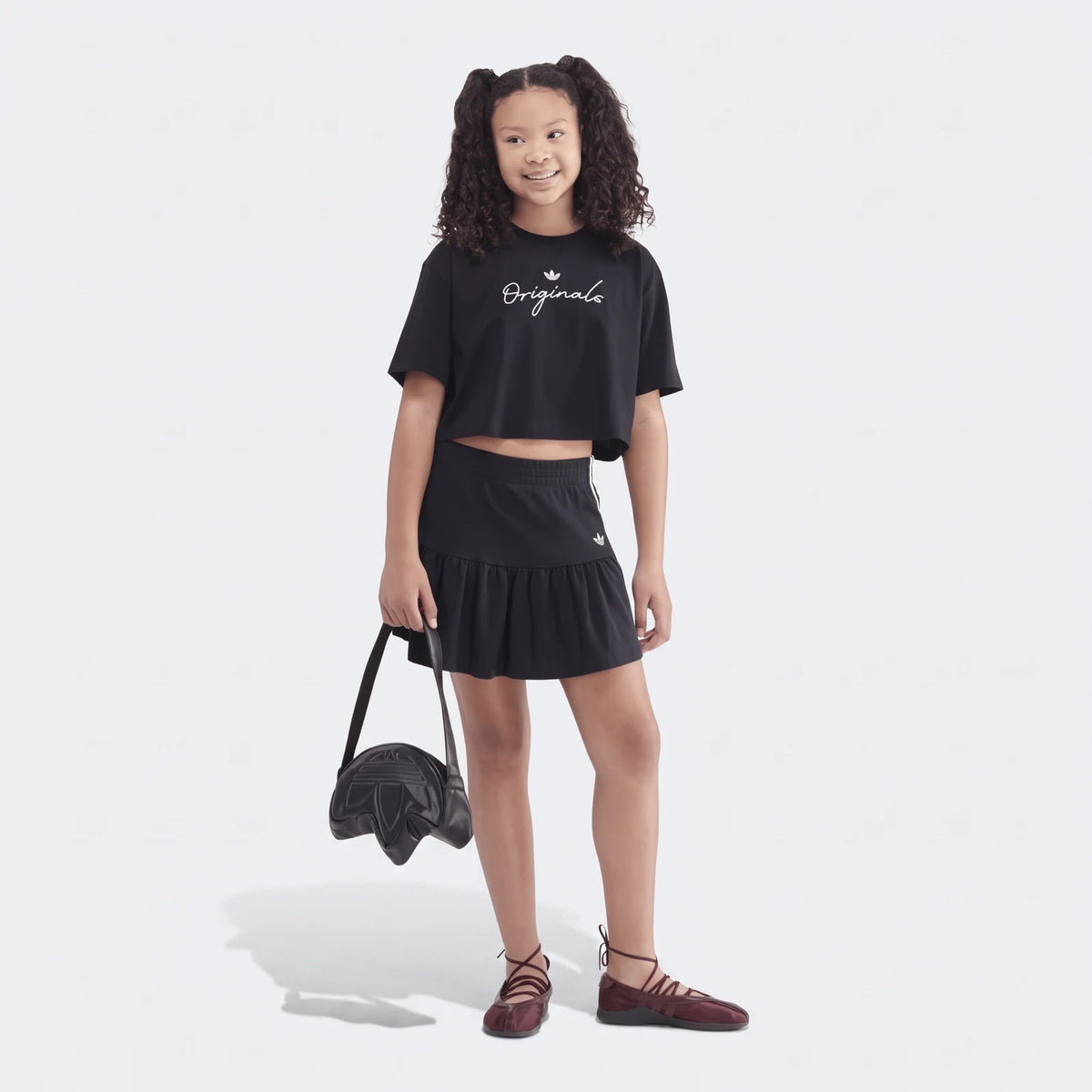 Young girl wearing a black Youth Script Tee and matching ruffled skirt, holding a black Adidas Trefoil bag with burgundy