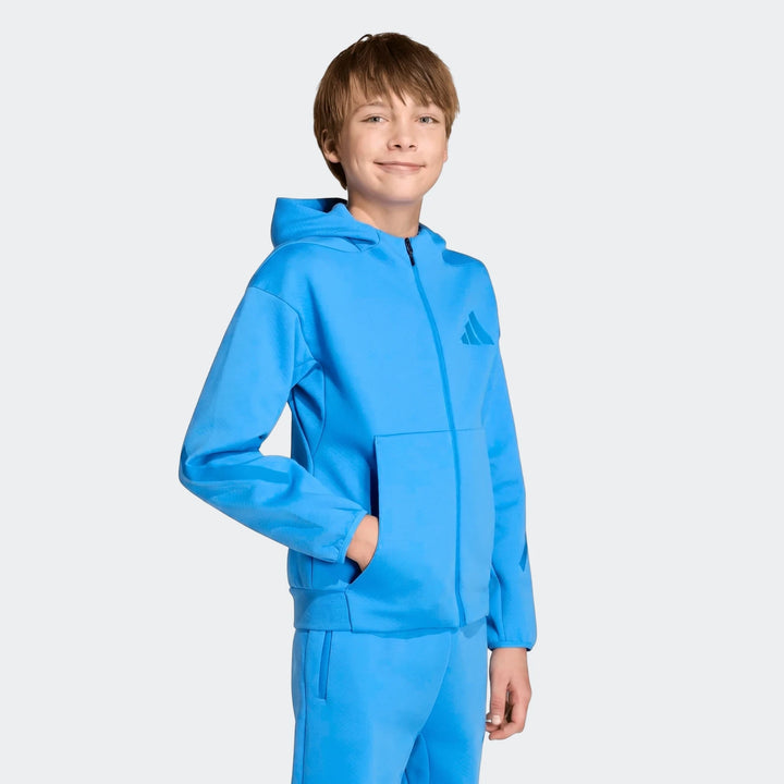 A young boy smiles wearing a blue Youth Z.N.E. Full-Zip Hoodie and matching sweatpants, posing casually with his hand in the