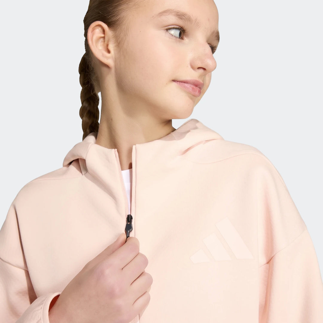A young girl in a light pink Adidas Youth Z.N.E. Full-Zip Hoodie, zipping up the front. The hoodie features a prominent