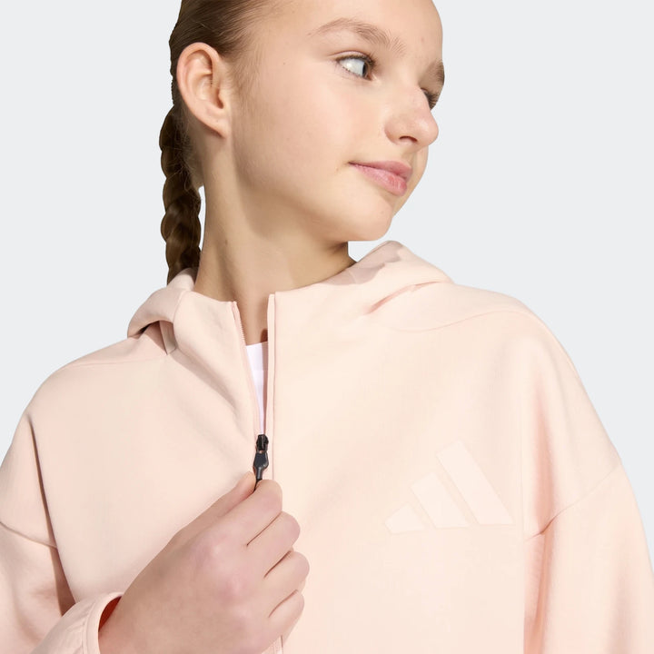 A young girl in a light pink Adidas Youth Z.N.E. Full-Zip Hoodie, zipping up the front. The hoodie features a prominent
