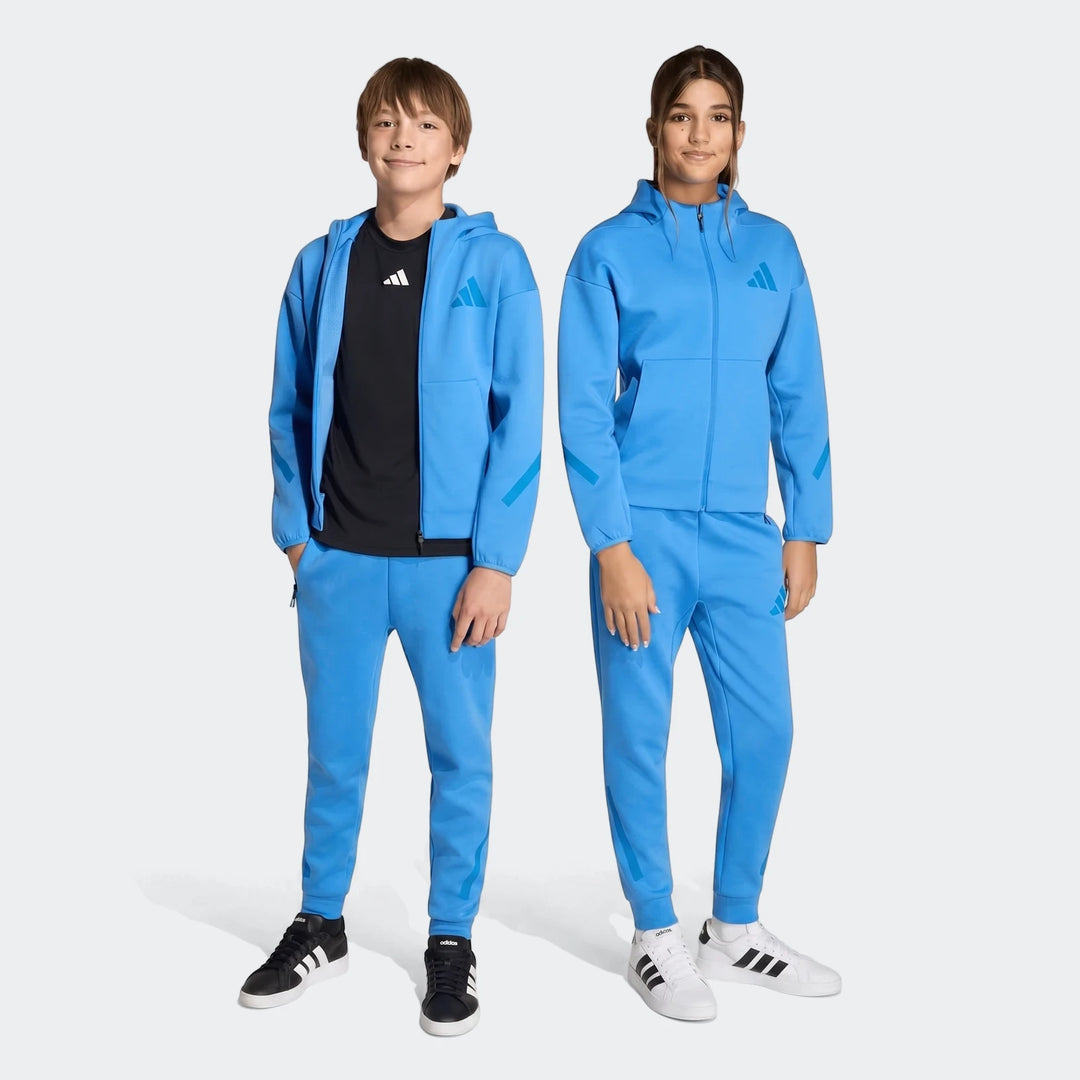 Two youth models wearing full blue Adidas Z.N.E. track pants and matching hooded jackets with black and white sneakers.