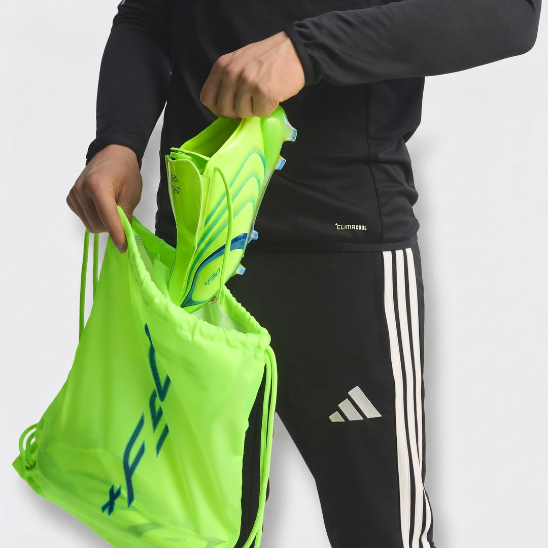 Person in black Z.N.E. Track Top and Adidas pants putting neon green soccer cleats into a matching drawstring bag.